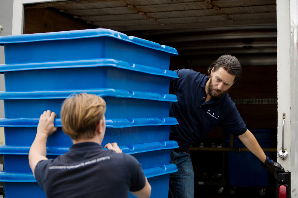 Two men moving large blue commercial moving bins that say DLO on the sides